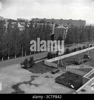 Ein Panorama-Hochwinkel-Archivfoto aus den 1980er Jahren, das die weitläufige urbane Landschaft des Artema-Mikrobezirks in Sloviansk, ukrainische SSR, UdSSR, erfasst. Das Schwarz-weiß-Bild zeigt eine riesige Stadtlandschaft, die von Reihen typischer Plattenwohnungen aus der Sowjetzeit („Panelkas“) dominiert wird. Im Vordergrund entfaltet sich eine Straßenszene mit einem umzäunten Hof mit einem Oldtimer und einem LKW. Dieser eindrucksvolle Blick verkörpert perfekt die Atmosphäre und den Maßstab einer Provinzstadt während der späten Sowjetzeit, bekannt als Perestroika. Stockfoto