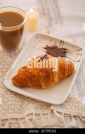 Frühstücksszene mit Croissant und Kaffee. Frühstück mit einem goldenen Croissant auf einem dekorativen Teller mit Kaffee und einem weichen Kerzenschein-Glo Stockfoto