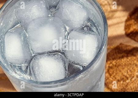 Eiswürfel in einem Glas Wasser auf einer gewebten Matte. Nahaufnahme eines klaren Glases gefüllt mit mehreren großen Eiswürfeln und Wasser. Selektiver Fokus. Stockfoto