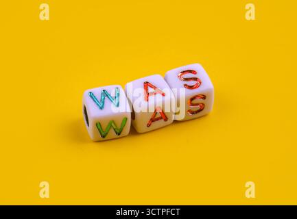 Drei weiße Würfel mit farbenfrohen Gravurbuchstaben, die das Wort „WAS“ schreiben, auf einem hellgelben Hintergrund platziert. Die verspielten Würfel symbolisieren Sprache. Stockfoto