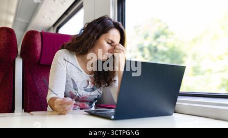 Junge kaukasische Frau im Zug, die sich müde fühlt, während sie einen Laptop benutzt Stockfoto