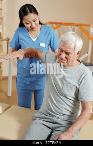 Physiotherapeut, der mit dem Patienten arbeitet. Ein alter kaukasischer Mann lächelt während er im Erholungszentrum körperliche Übungen mit einer jungen, pflegenden asiatischen Krankenschwester macht Stockfoto