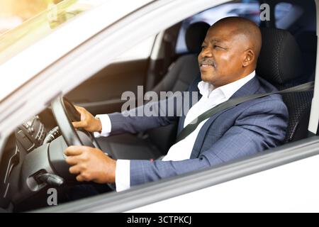 Lächelnder afroamerikanischer Geschäftsmann, der Auto fährt, eleganten Anzug und Sicherheitsgurt trägt Stockfoto