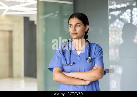 Frau im Gesundheitswesen, die blaue Peelings und ein Stethoskop trägt, mit überkreuzten Armen steht und wegblickt, repräsentiert Hingabe und kritisches Denken in ihrer medizinischen Rolle Stockfoto