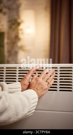 Die Hände wärmen sich in der Nähe eines elektrischen Konvektors in einem sanft beleuchteten Wohnzimmer Stockfoto