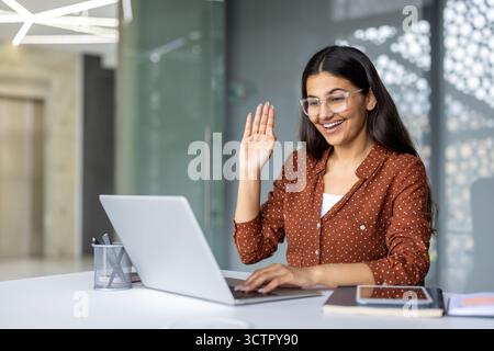 Junge indische Geschäftsfrau, die in einem modernen Büro auf einem Laptop eine Videokonferenz anruft, lächelnd und winkend mit der Hand, die Fernarbeit und Online-Kommunikation zeigt Stockfoto