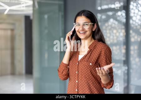 Junge indische Profi-Frau mit Brille steht in einem modernen Büro, lächelnd und animiert, während sie auf ihrem Smartphone spricht, und vermittelt ein selbstbewusstes, freundliches Geschäftsgespräch Stockfoto