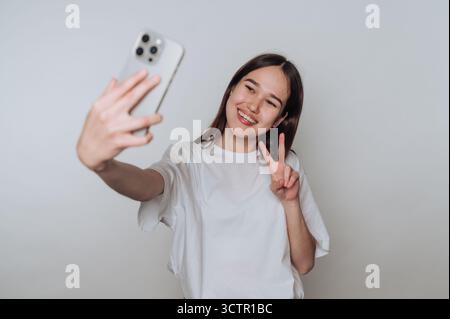 Junge Frau, die ein Selfie mit einem Smartphone macht, lächelt und ein Friedenszeichen vor einem einfachen Hintergrund zeigt. Stockfoto