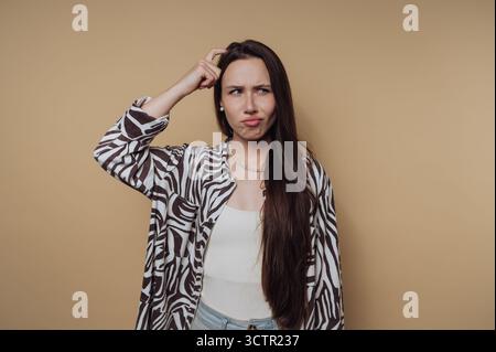 Frau kratzt sich verwirrt den Kopf vor beigefarbenem Hintergrund. Stockfoto