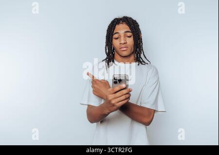 Junger Mann mit geflochtenen Haaren, der auf das Smartphone zeigt, neutraler Hintergrund. Stockfoto