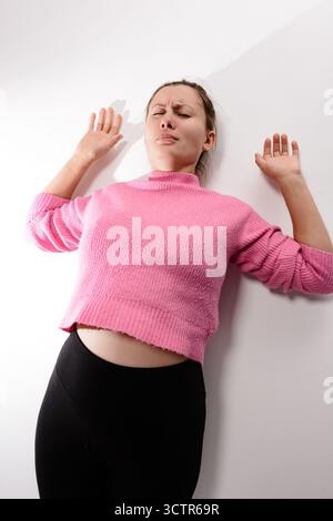 Frau in rosafarbenem Oberteil und schwarzer Leggings lehnt sich mit geschlossenen Augen und schmerzhaftem Ausdruck an die weiße Wand. Stockfoto