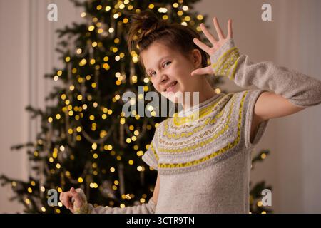 Lächelndes Teenager-Mädchen in gemütlichem Strickkleid posiert verspielt mit erhobener Hand und zeigt fünf Finger neben einem verzierten Weihnachtsbaum mit festlichem Licht Stockfoto