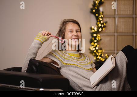 Teenager im gestrickten Winterkleid lächelt und spielt mit Zuckerrohr, während sie sich auf einem schwarzen Ledersofa mit offenem Notizbuch und festlichem Weihnachtslicht entspannt Stockfoto