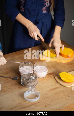 Nahaufnahme von Kinderhänden, die ein gesundes Dessert mit frischer Mango, Pflanzenmilch und Chiasamen auf einem Holztisch vorbereiten, ein Kind schneidet das Mangoweiß Stockfoto
