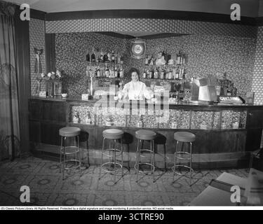 Barkeeperin in den 1960ern Eine Frau steht hinter der Bar, die Regale mit Flaschen an der Wand dahinter hat. Sie hält zwei Getränke in der Hand, die sie zubereitet hat. 1960 Kristoffersson Ref. 356A-16 Stockfoto