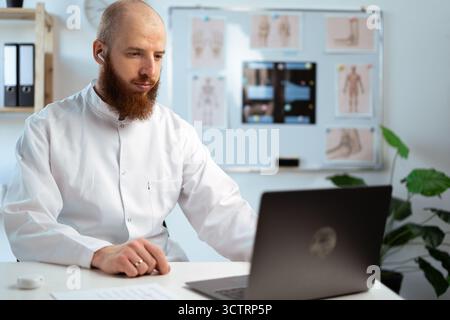 Bärtiger männlicher Arzt mit weißem Mantel im Krankenhausbüro mit Laptop-Computer, Analyse medizinischer Daten. Stockfoto