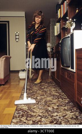 So sah ein typisches Familienhaus in den 1970er Jahren aus Eine Frau, die Clogs trägt, saugt den Teppich im Wohnzimmer ab. Ein großes braunes Bücherregal ist zu sehen, zusammen mit einem für die Zeit typischen Fernseher in einem weißen Schrank. Schweden, 1979. Kristoffersson Ref. CV37-17. Stockfoto
