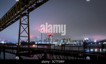 Die Skyline der Londoner Docklands bei Nacht mit Kränen und einem industriellen Fußweg über den Fluss Stockfoto