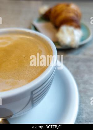 Nahaufnahme einer Tasse Kaffee mit Schaum und einem verschwommenen Croissant mit Sahne, ideal für ein morgendliches Essen Stockfoto