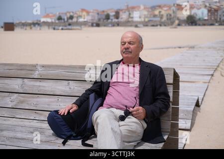 Ein älterer Mann sitzt auf einer Bank am Strand, ruht sich aus und sonnt sich Stockfoto