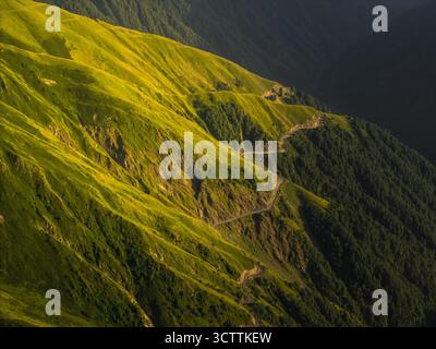 Aerial view of a winding road snakes through vibrant green mountains contrasting with deep shadows, Alazani pass, Chala, Kakheti, Georgia. Stockfoto