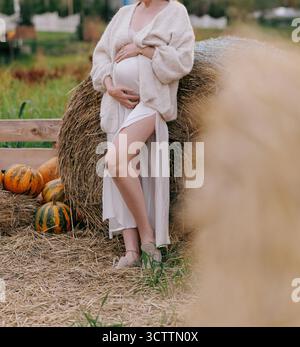 Schöne schwangere Frau, die den Bauch während des Herbstes berührt, Foto-Shooting ländliche Lage vor dem Hintergrund von Heuballen und Kürbissen. Konzept Happy m Stockfoto