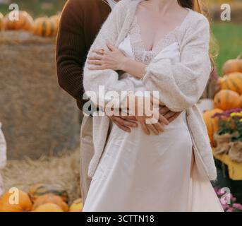 Glückliches schwangeres Paar umarmt sich mit Liebe und Sorgfalt, Mann hält den Babybump der Frau während Outdoor-Herbstfoto-Shooting mit Kürbissen. Konzept Familienerwartung Stockfoto