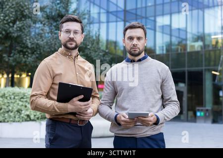 Zwei selbstbewusste Männer in Business-Freizeitkleidung, die ernsthaft mit einem Ordner und einem digitalen Tablet in einer modernen urbanen Umgebung stehen und für Partnerschaft, Teamwork und Planung stehen Stockfoto