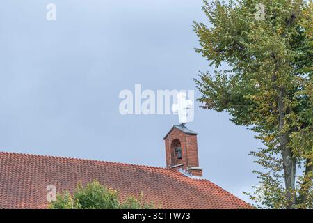 Dach einer katholischen Kirche mit einem großen weißen Kreuz vor blauem Himmel und grauen Wolken, hohe grüne Bäume in der Nähe Stockfoto