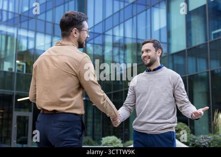 Zwei lächelnde Erwachsene Männer schütteln sich die Hände und treten miteinander zusammen, um vor einem modernen Bürogebäude einen erfolgreichen Geschäftsabschluss oder eine Vereinbarung abzuschließen Stockfoto