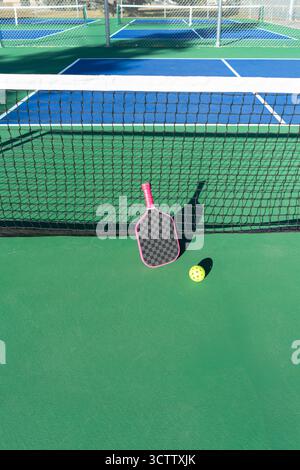 Niedriger Winkel des Pickleball-Paddels, das gegen das Netz eines Pickleball-Platzes im Freien gelehnt ist, grünes und blaues Spielfeld Stockfoto