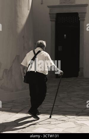 Schwarz-weiß-Bild einer traditionellen Szene in der Altstadt von naxos, als ein älterer grieche mit Gehstock allein zu einem kunstvollen Eingang geht Stockfoto