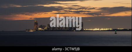 Weit entfernte Skyline von Forest City, Johor, Malaysia, von der Westküste Singapurs in der Abenddämmerung aus gesehen. Beleuchtete Gebäude und Hafenkräne sichtbar. Stockfoto