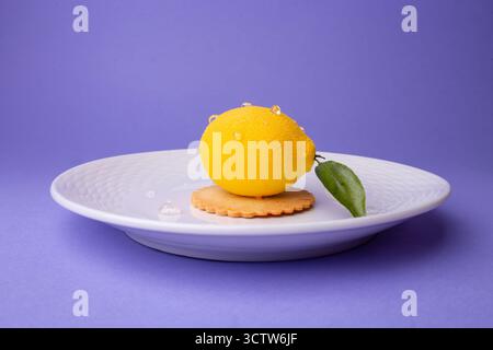 Seitliche Aufnahme eines zitronenförmigen Desserts: Strukturierte Oberfläche, glitzernde Wassertröpfchen, grünes Blatt und knusprige Basis auf einem weißen gerippten Teller. Stockfoto