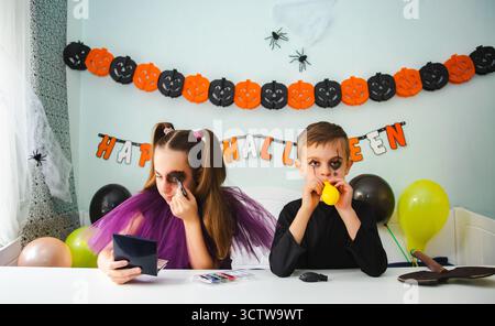 Zwei Kinder bereiten sich auf Halloween vor. Ein Mädchen schminkt sich, während der Junge den Ballon aufbläht. Dekorierter Hintergrund mit Kürbissen und Bannern. Stockfoto