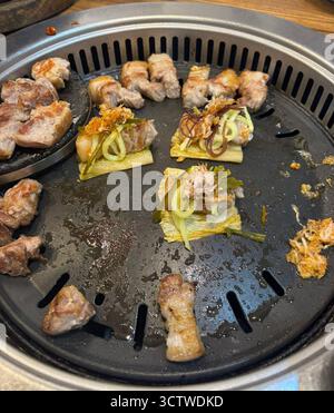 Ein Foto von gegrilltem Schweinebauch und verschiedenen gewickelten Fleischgerichten, serviert auf einem Grill in einem Restaurant im Vorort Busan, Südkorea. Koreanisches Barbecue (K-BBQ) ist ein sehr geselliger Brauch, bei dem die Gäste verschiedene Fleischstücke, oft Schweine- oder Rindfleisch, auf einem Grill kochen, der in ihren Tisch eingelassen ist. Das gekochte Fleisch wird häufig in Salat oder anderem Blattgemüse (Sam) gewickelt, oft begleitet von Reis, Knoblauch, Chili und verschiedenen eingelegten Beilagen (Banchan) und Saucen. 2025. Stockfoto
