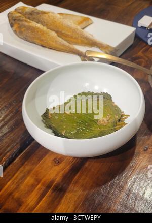 Ein Foto der traditionellen koreanischen Küche in Gyeongju, Südkorea, mit gewürzten Perillablättern in einer weißen Schüssel und in der Pfanne gebratenem ganzer Fisch auf einer rechteckigen Platte. Die koreanische Küche ist eine reiche kulinarische Tradition, die von der landwirtschaftlichen Geschichte, dem Klima und dem buddhistischen Erbe des Landes beeinflusst wird, mit Banchan (??) oder Beilagen, integraler Bestandteil der Mahlzeiten. 2025. Stockfoto