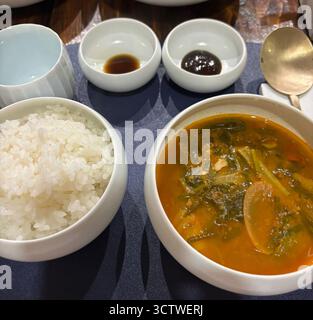 Ein Foto der traditionellen koreanischen Küche in Gyeongju, Südkorea, mit einer Schüssel flauschigen weißen Reis, einem leuchtenden orange-roten Eintopf mit grünem Blattgemüse und zwei kleinen Schüsseln dunkler Saucen. Traditionelle koreanische Küche, oft auch als ??? Bezeichnet (Hanjeongsik), zeichnet sich durch eine Vielzahl von Beilagen (??, Banchan) aus, die neben Reis und Suppe oder Eintopf serviert werden. 2025. Stockfoto