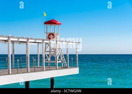 Rettungsschwimmerstuhl am Ende eines Piers über dem Strand in einem Badeort an einem sonnigen Tag. Ruhiges Meer und blauer Himmel. Konzept des Badeurlaubs. Keine Personen Stockfoto