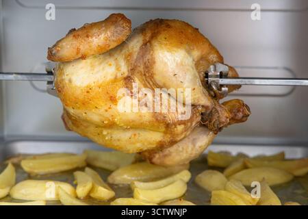 Saftiges, goldgeröstetes Huhn auf einem Spieß über einem Tablett mit gewürzten Kartoffelscheiben in einem warmen, heimeligen Ofen, einladend für ein entspannendes Familienessen. Stockfoto