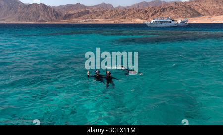 Dahab, Ägypten - 5. Februar 2024: Drei Personen schnorcheln im türkisfarbenen Wasser des Roten Meeres mit Bergen, Yachten und der Stadt Dahab im Hintergrund Stockfoto