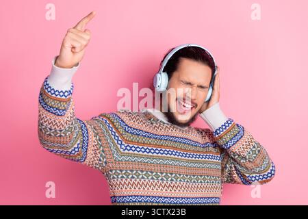 Fröhlicher Mann mit gemustertem Pullover, Musik auf Kopfhörern hören und leidenschaftlich vor leuchtendem rosa Hintergrund singen Stockfoto