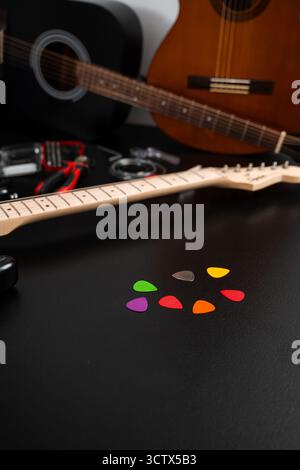 Gitarrenpicks mit Gitarren und Werkzeugen im Hintergrund. Eine Reihe von bunten Gitarrenpicks liegt auf einer schwarzen Oberfläche mit Gitarren und Reparaturwerkzeugen. Mehrere Gitarren Stockfoto