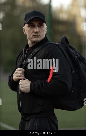 Ein Mann, der eine schwarze Bomberjacke und -Mütze trägt, posiert selbstbewusst mit einem Rucksack über der Schulter in einem grünen Park an einem klaren Tag. Bäume sind im Hintergrund sichtbar. Stockfoto