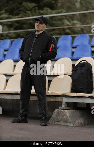 Ein Mann mit einer schwarzen Bomberjacke und -Mütze steht in den leeren Ständen und konzentriert sich auf das Spiel unter ihm. Seine Körpersprache zeigt Vorfreude und Engagement für die Leistung des Teams. Stockfoto