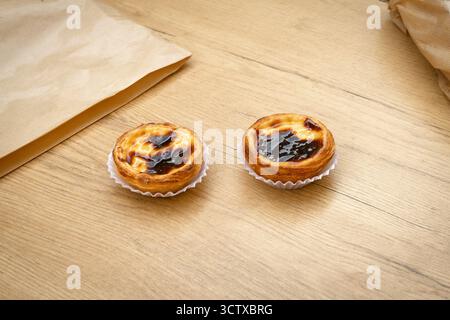 Zwei perfekt karamellisierte Pastéis de Nata (portugiesische Pudding-Torten) auf einem strukturierten Holztisch neben zerknitterten Papiertüten. Süßer Snack Stockfoto
