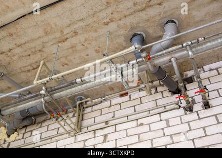 Überhängende industrielle Sanitär- und Heizungssysteme mit einem Netz aus Metallwasserleitungen, Armaturen und Kunststoffabflussleitungen unter einer Fabrikdecke. Stockfoto