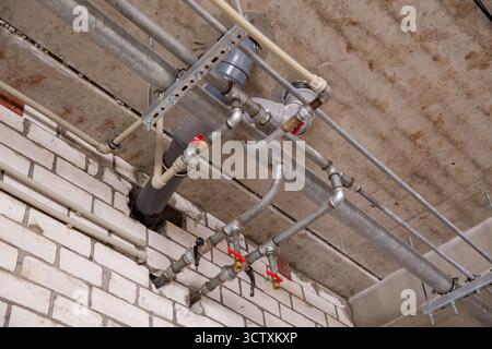 Überhängende industrielle Sanitär- und Heizungssysteme mit einem Netz aus Metallwasserleitungen, Armaturen und Kunststoffabflussleitungen unter einer Fabrikdecke. Stockfoto