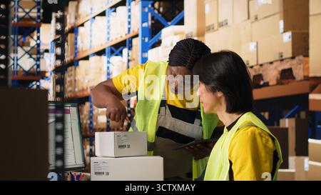 Zwei Kollegen arbeiten an der Auftragsbearbeitung in industriellen Lagerhäusern und tragen Warnwesten in der Nähe von riesigen Lagerregalen. Asiatischer Mann, der die Versanddaten auf dem Computer eingibt. Kamera B.. Stockfoto