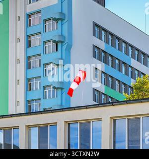 Rote und weiße Windsocken flattern im Wind auf dem Dach des modernen Gebäudes mit blauer und grüner Fassade. Windsocken-Konzept, städtische Windrichtung, Wetter Stockfoto